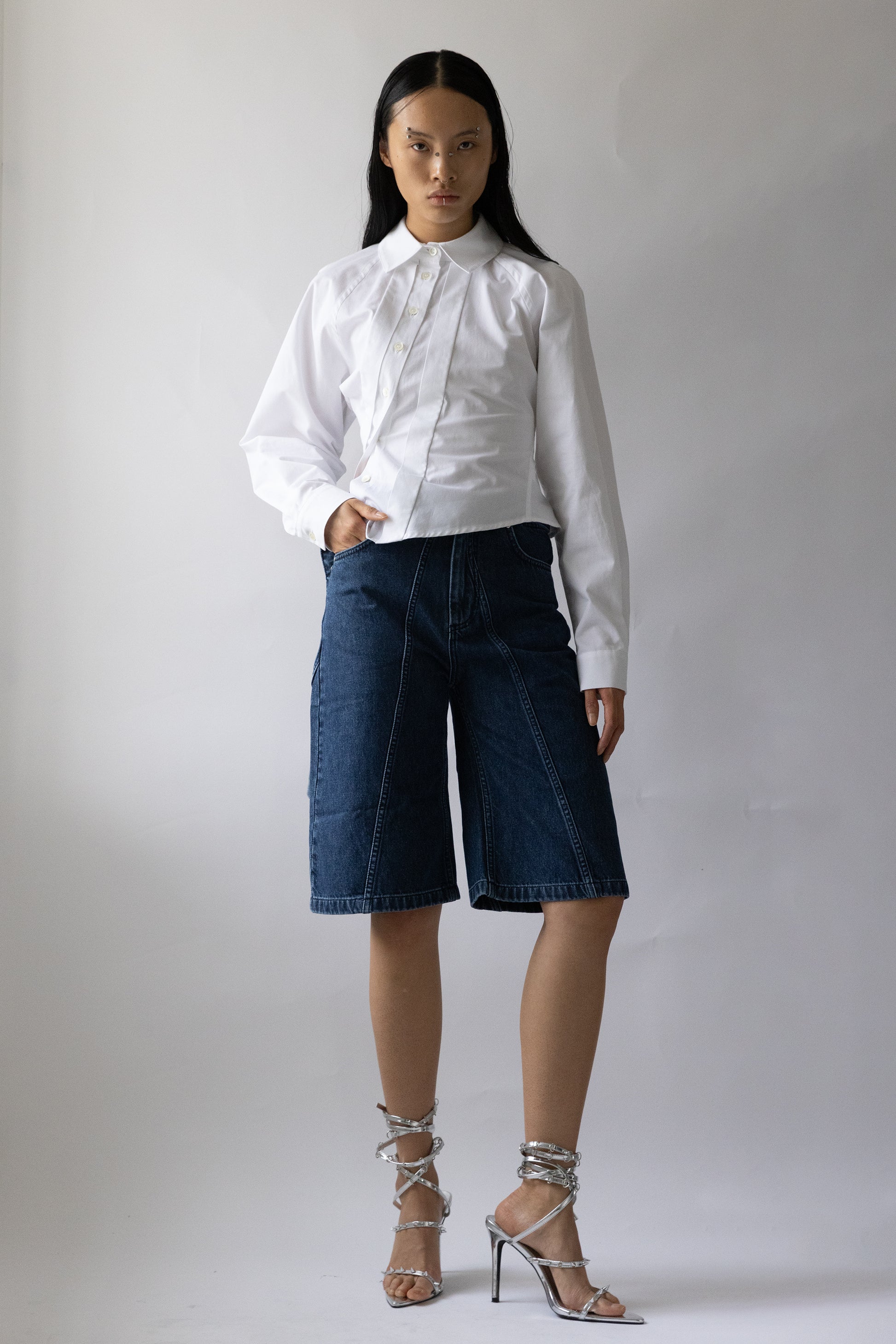 model front view wears knee length blue denim garment with five pockets and raw hem by Jade Cropper